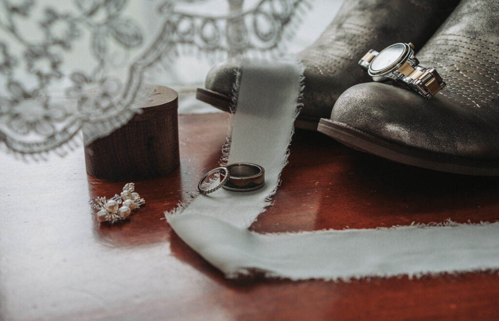 Close-up of wedding accessories on a wooden table, including groom's shoes, a watch, wedding rings, and a lace veil. Wedding at the Walnut Valley House shot by Quinn Photography LLC