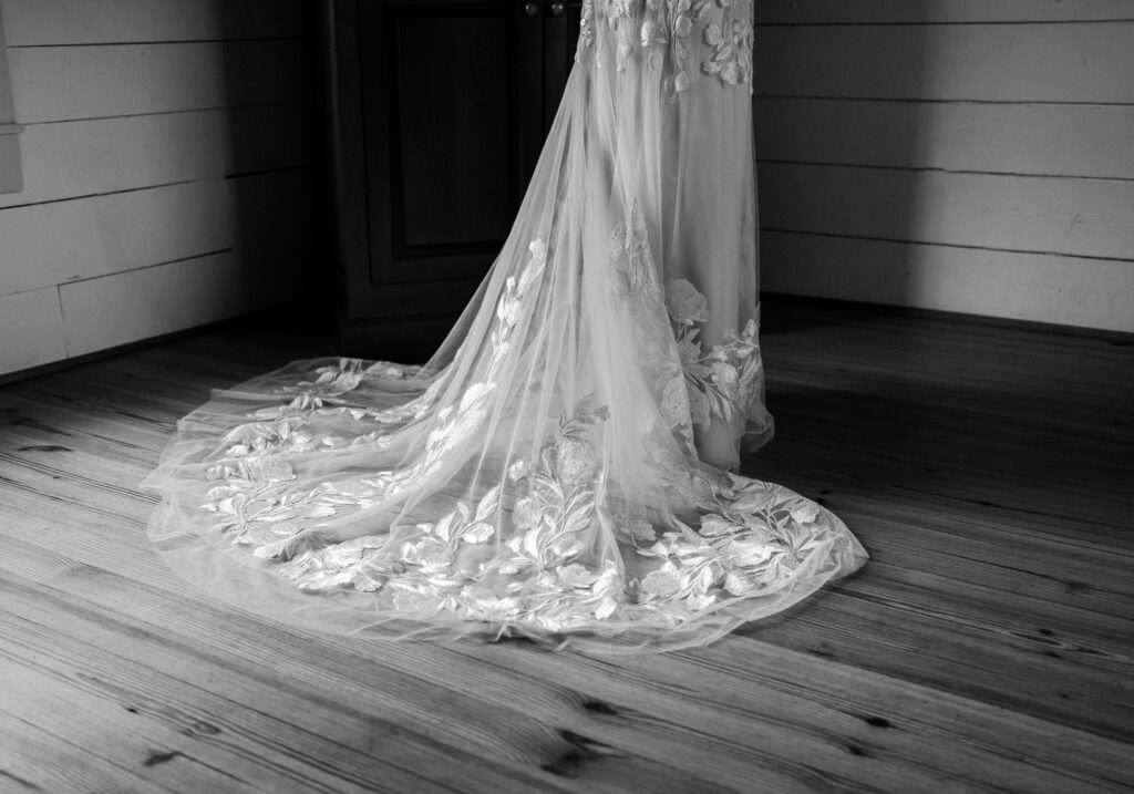 Black and white image of a bridal gown's detailed lace train spread elegantly on a wooden floor, highlighting its intricate floral embroidery. Quinn Photography LLC