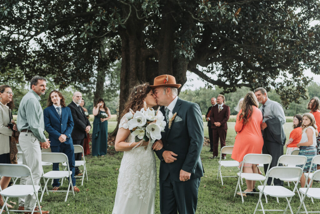 A bride and groom kissing passionately under a large tree during their wedding ceremony, surrounded by guests in a variety of colorful attire. Wedding at the Walnut Valley House shot by Quinn Photography LLC