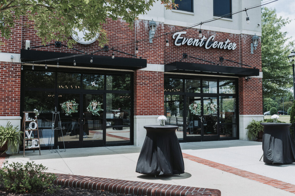 Alt Description: Entrance of a modern event center with a brick facade, featuring large windows and doors framed by romantic floral arrangements and a "LOVE" sign. Hampton Roads Event Center. Quinn Photography LLC