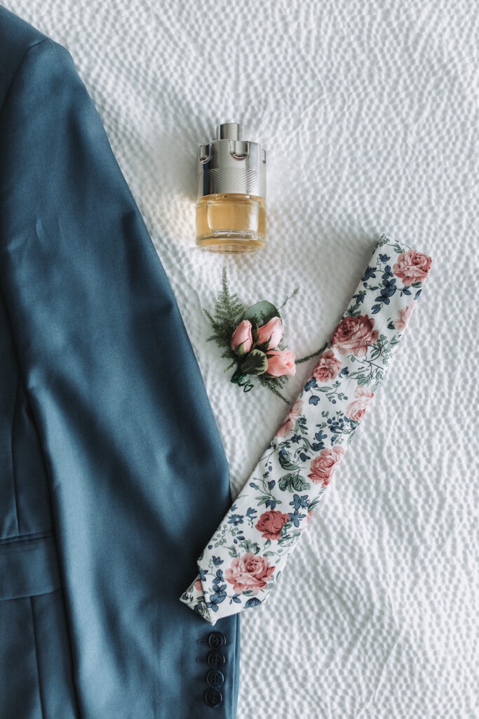 A groom's wedding day accessories laid out on a textured white bedspread: a navy suit jacket, a floral patterned tie, a boutonniere with pink roses, and a bottle of cologne.