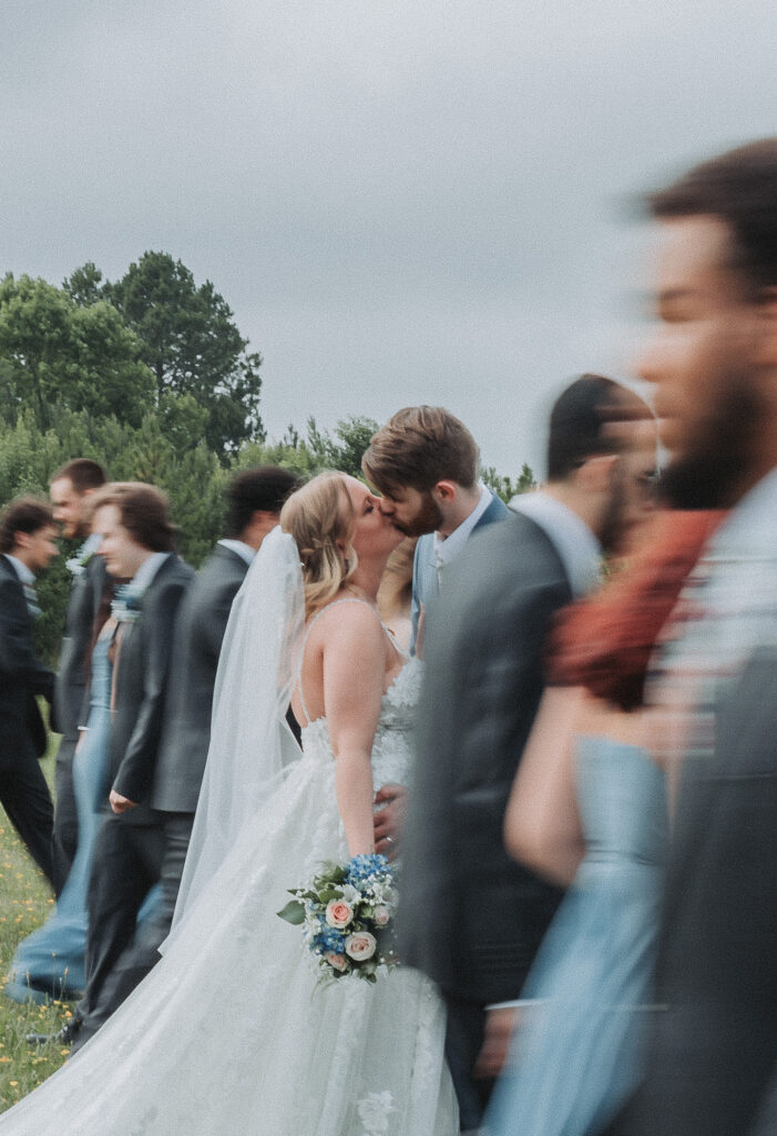 In a dynamic, blurred scene, a bride and groom share a kiss amidst their wedding guests, focusing sharply on their intimate moment. Quinn Photography LLC