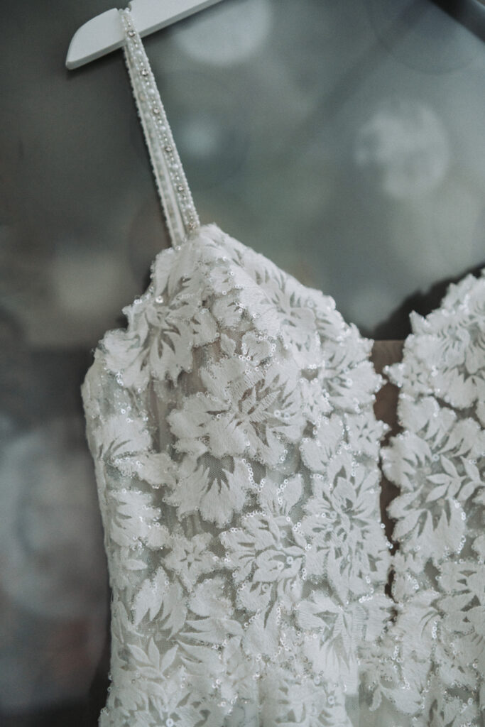 Close-up of a bridal gown's intricate lace detail and beaded embellishments, hanging from a pearl-studded hanger. Quinn Photography LLC