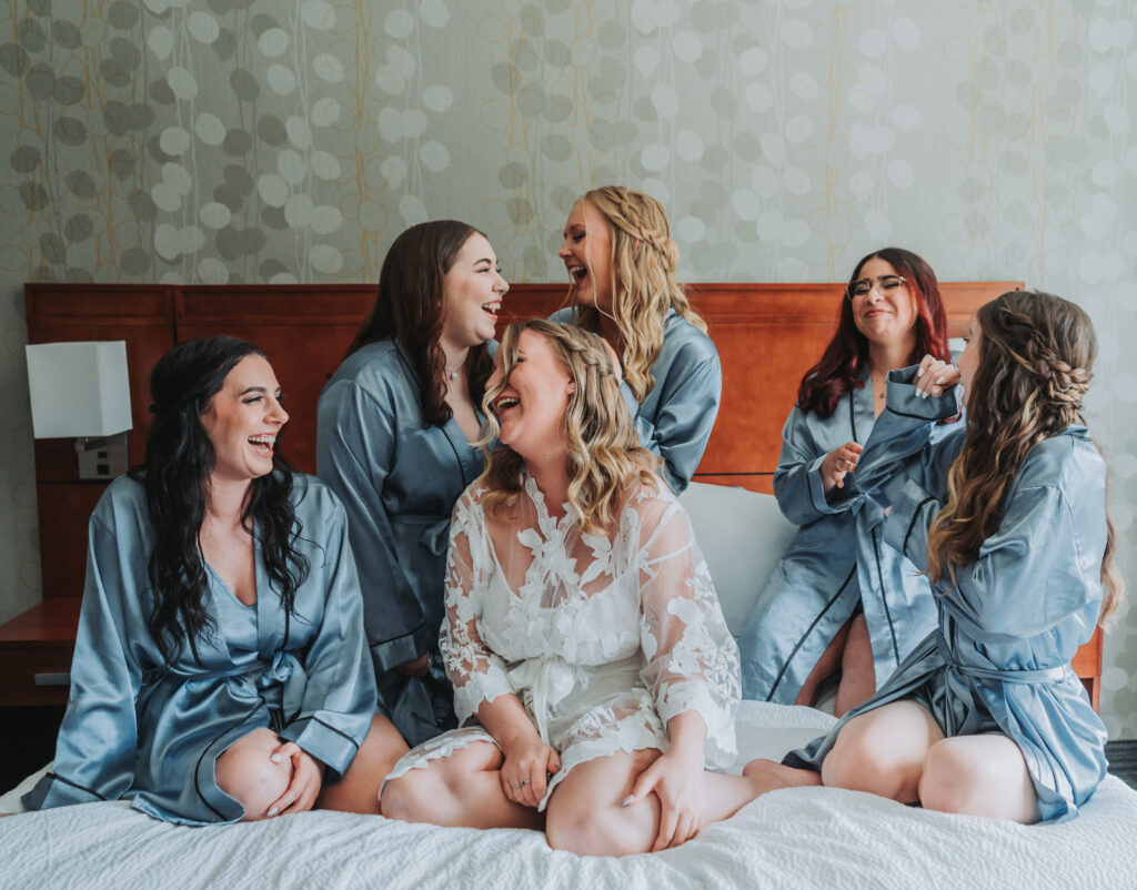 A bride in a white lace robe laughs joyously with her bridesmaids, who are all dressed in matching blue silk robes, in a hotel room.