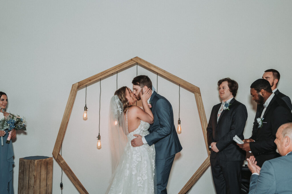 A bride and groom share their first kiss as a married couple, framed by a geometric wooden arch with hanging Edison bulbs, while the bridal party watches with joy. Quinn Photography LLC