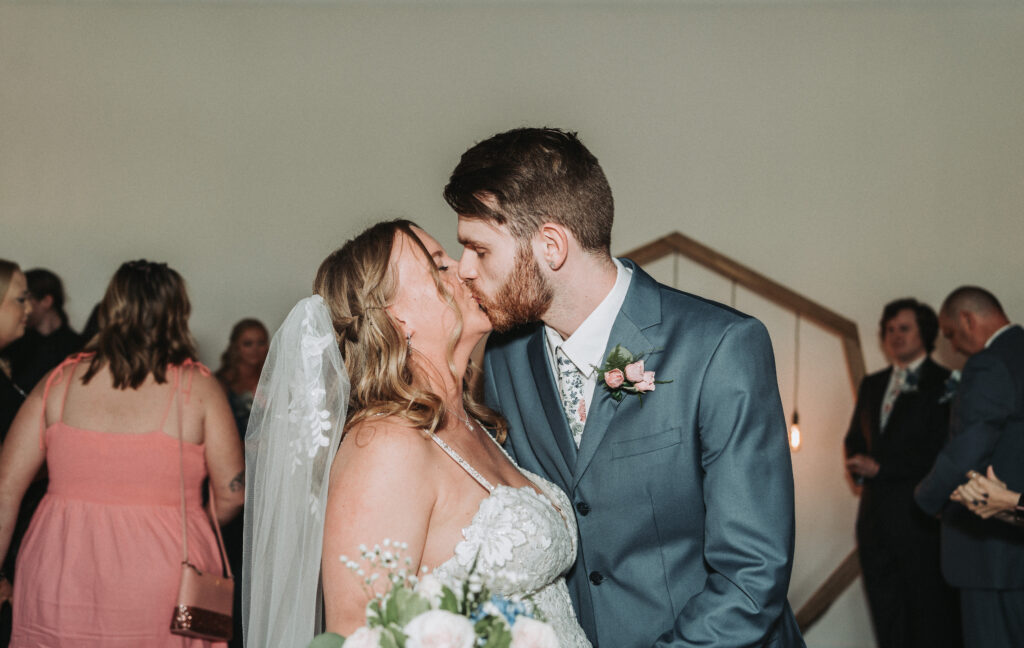 A bride and groom sharing a tender kiss at their wedding ceremony, with the bride's delicate lace veil and the groom's stylish blue suit in sharp focus. Quinn Photography LLC