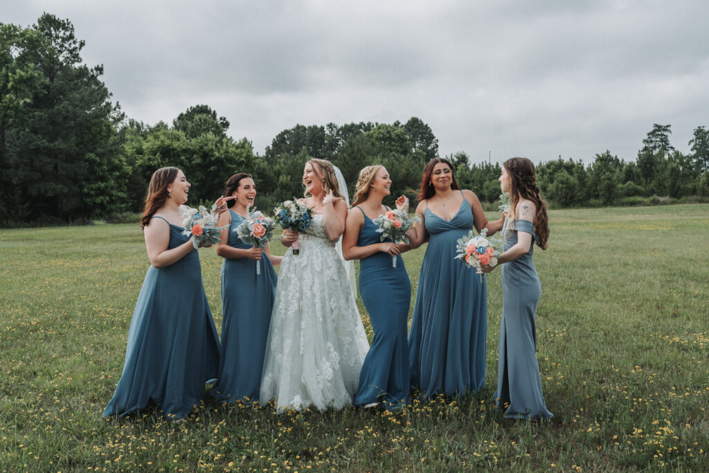 A joyful bride and her bridesmaids dressed in varying shades of blue, laughing together in a field dotted with wildflowers. Quinn Photography LLC