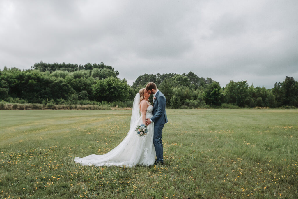 A bride and groom share a tender moment in a vast field dotted with wildflowers, under a cloudy sky. The bride's lace gown flows elegantly with a long train, as the groom, in a blue suit, embraces her gently. Quinn Photography LLC