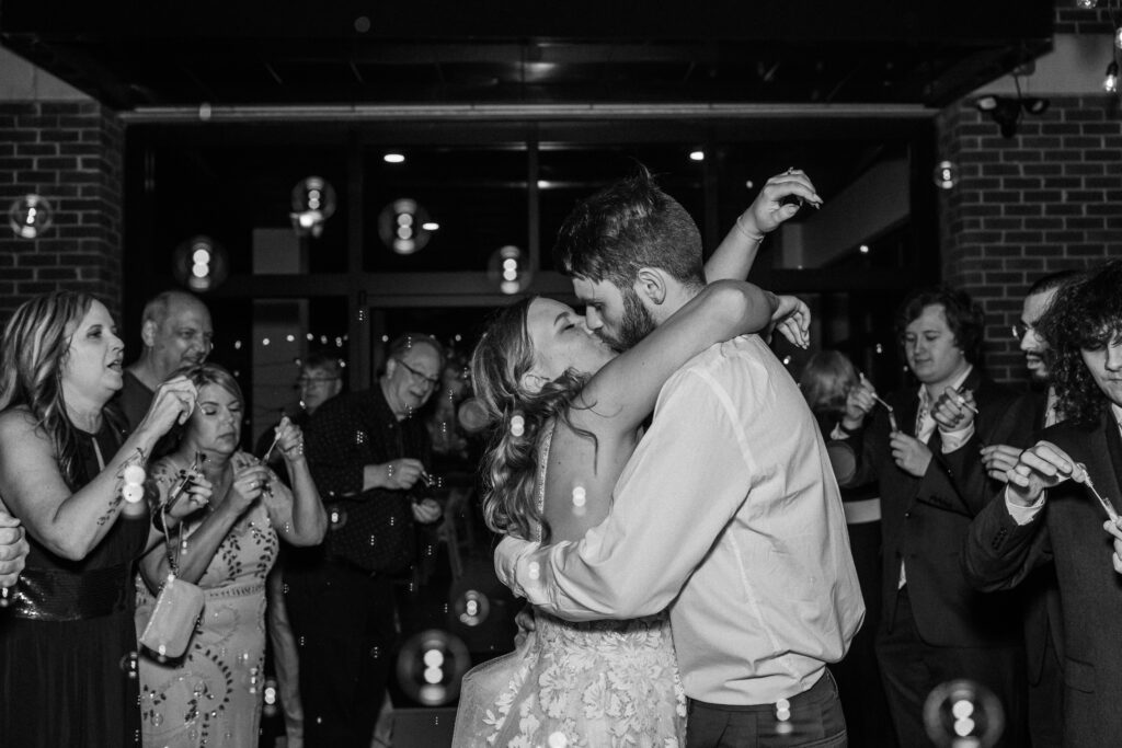 A black and white photo of a bride and groom sharing a passionate kiss during their bubble exit, surrounded by guests joyfully blowing bubbles. Quinn Photography LLC