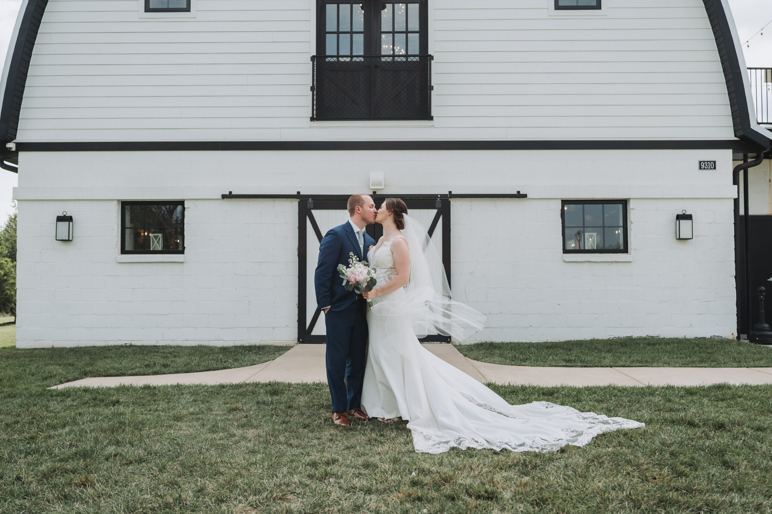 Bride and groom share a kiss outside Sweeney Barn in front of the white barn doors, with the bride’s veil flowing in the wind. Quinn Photography LLC