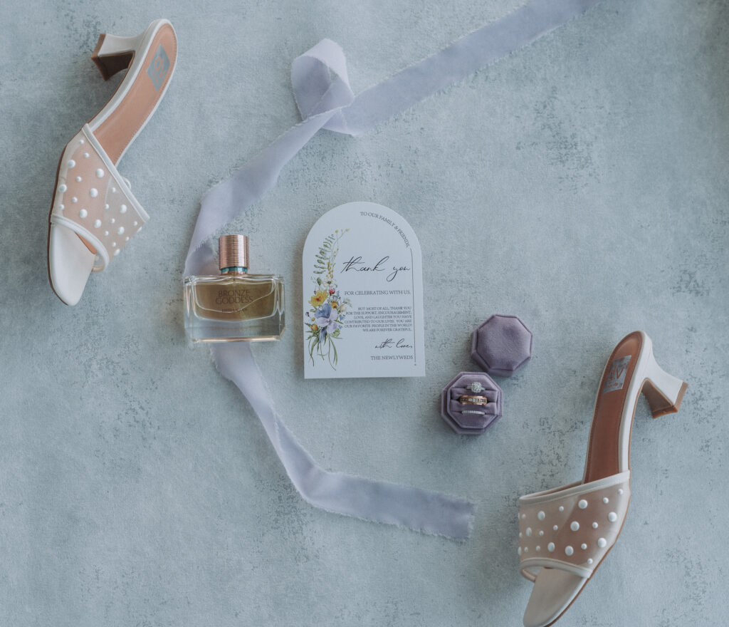 Bridal flat lay with pearl heels, Bronze Goddess perfume, wedding rings, purple velvet ring box, and thank-you card — styled and captured by Quinn Photography LLC.