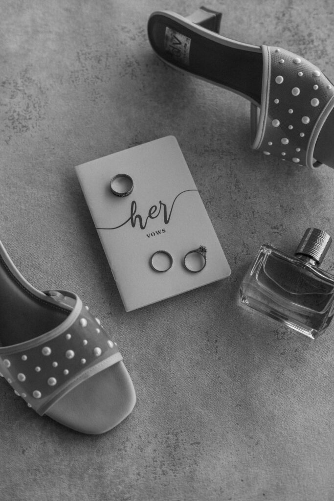 Black and white bridal flat lay with vow book, wedding rings, perfume, and pearl heels — photographed by Quinn Photography LLC.