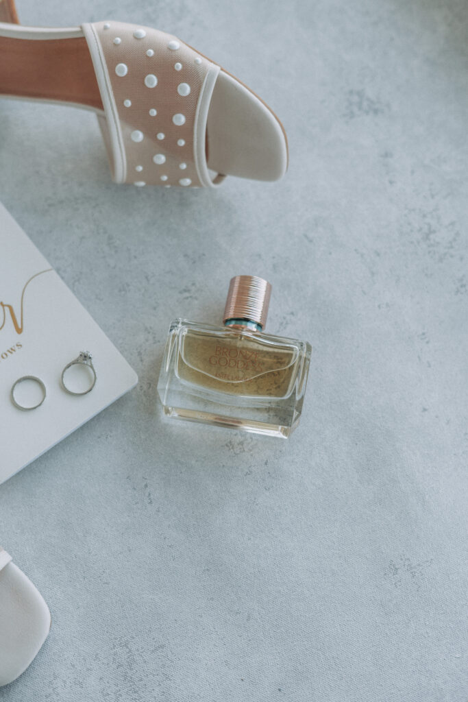Close-up of pearl bridal heel, Bronze Goddess perfume, and wedding rings near vow book — captured by Quinn Photography LLC.