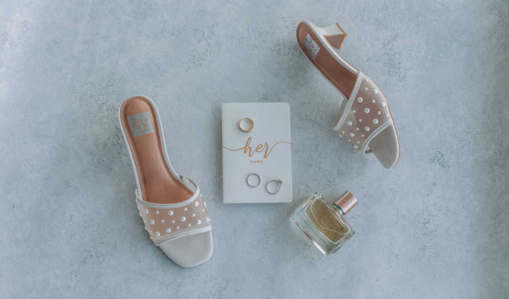 Top-down flat lay of vow book, pearl heels, perfume, and wedding rings on soft blue textured background — styled by Quinn Photography LLC.