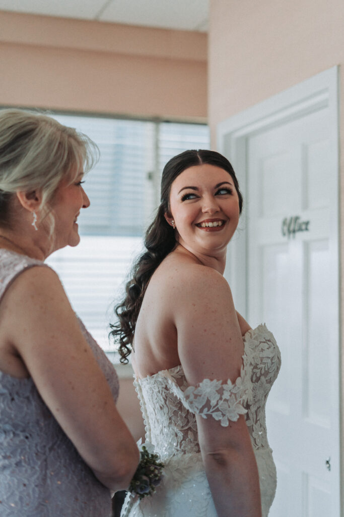 Bride smiling over her shoulder as her mother helps her get into her wedding dress at the Lesner Inn — photographed by Quinn Photography LLC.