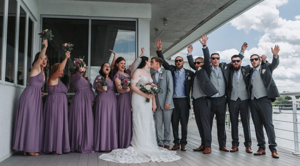 Bride and groom share a kiss surrounded by cheering bridal party in lavender and gray at Lesner Inn — photographed by Quinn Photography LLC.