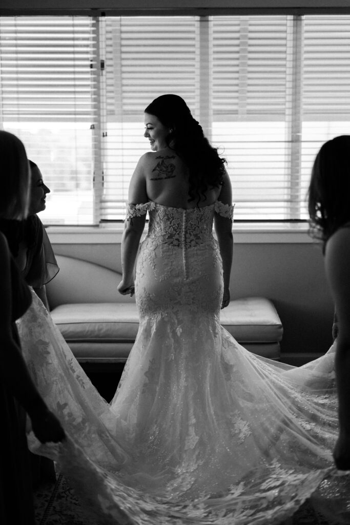 Black and white photo of bride smiling while bridesmaids adjust her lace train near a window — photographed by Quinn Photography LLC.