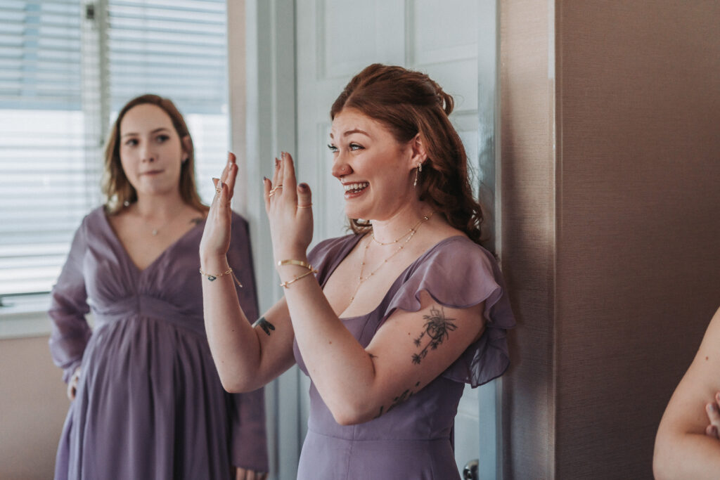 Bridesmaid in lavender dress reacts emotionally to seeing the bride, smiling with hands raised — captured by Quinn Photography LLC.