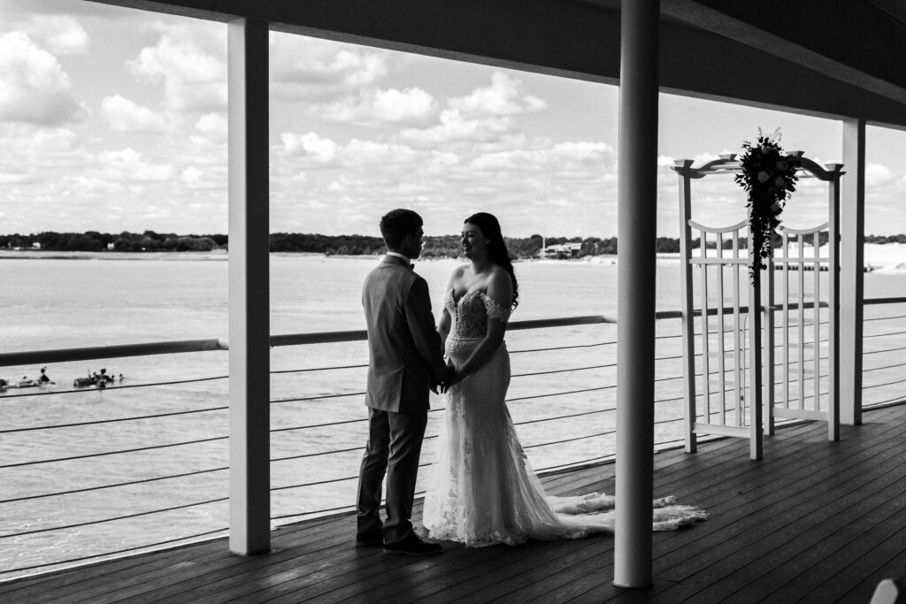 Bride and groom holding hands during first look, silhouetted against the water