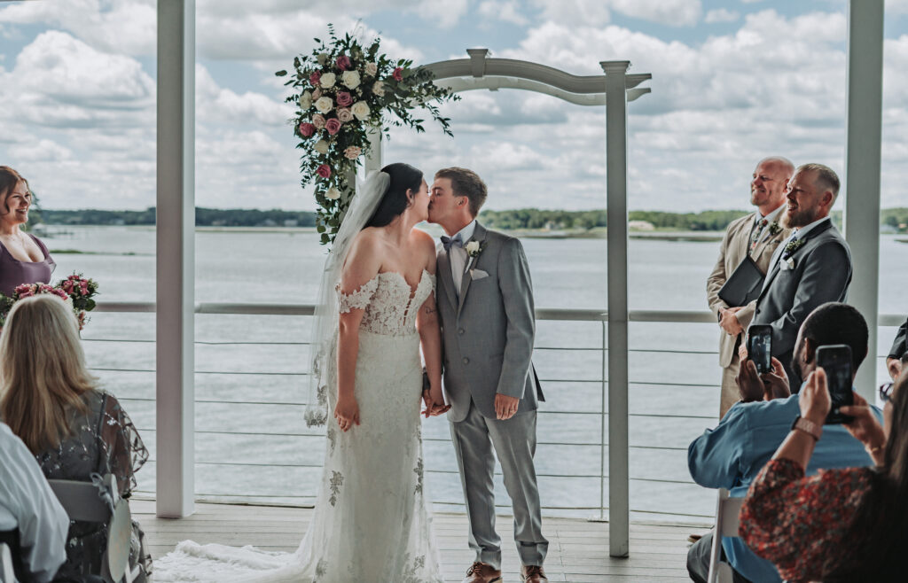 Bride and groom share a kiss under the floral ceremony arch by the water