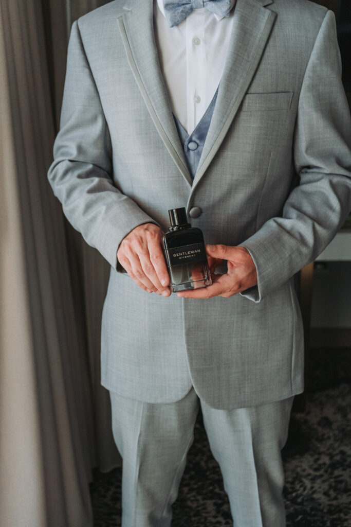Groom in light gray suit holding a bottle of Givenchy Gentleman cologne before the wedding ceremony.
