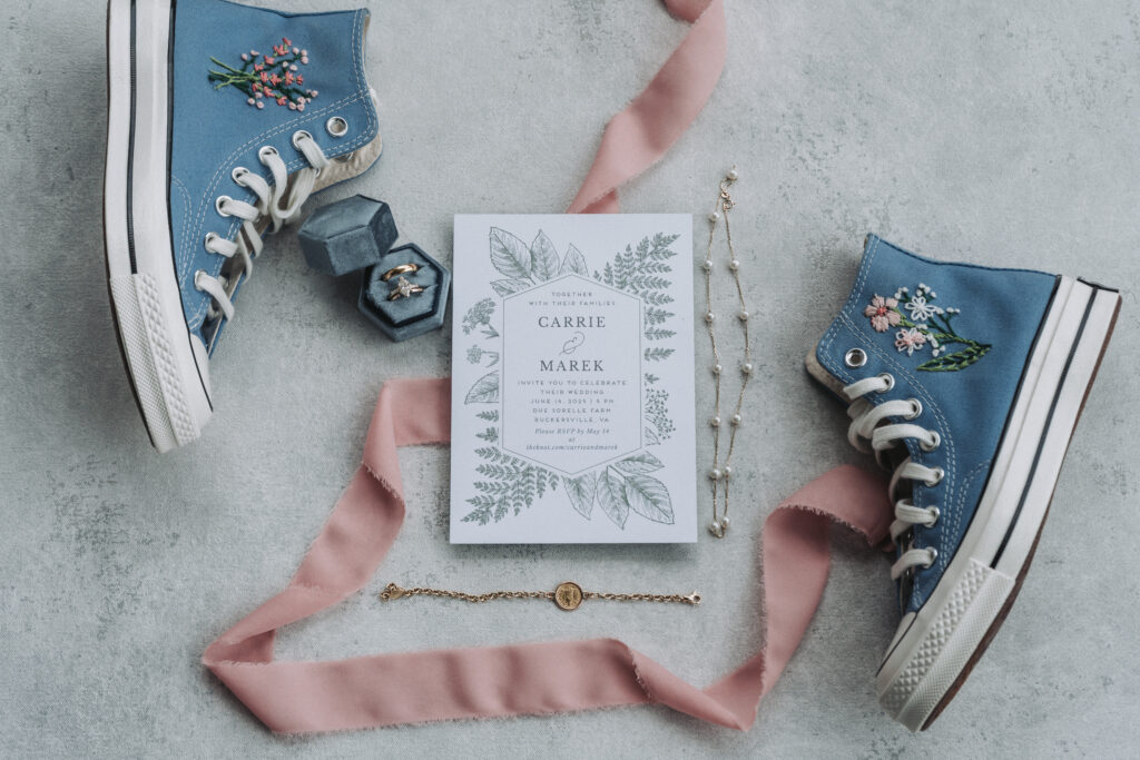 Wedding flatlay at Due Sorelle with embroidered blue Converse, velvet ring box, pink ribbon, and floral invitation. Captured by Quinn Photography LLC in Buckingham County, Virginia.