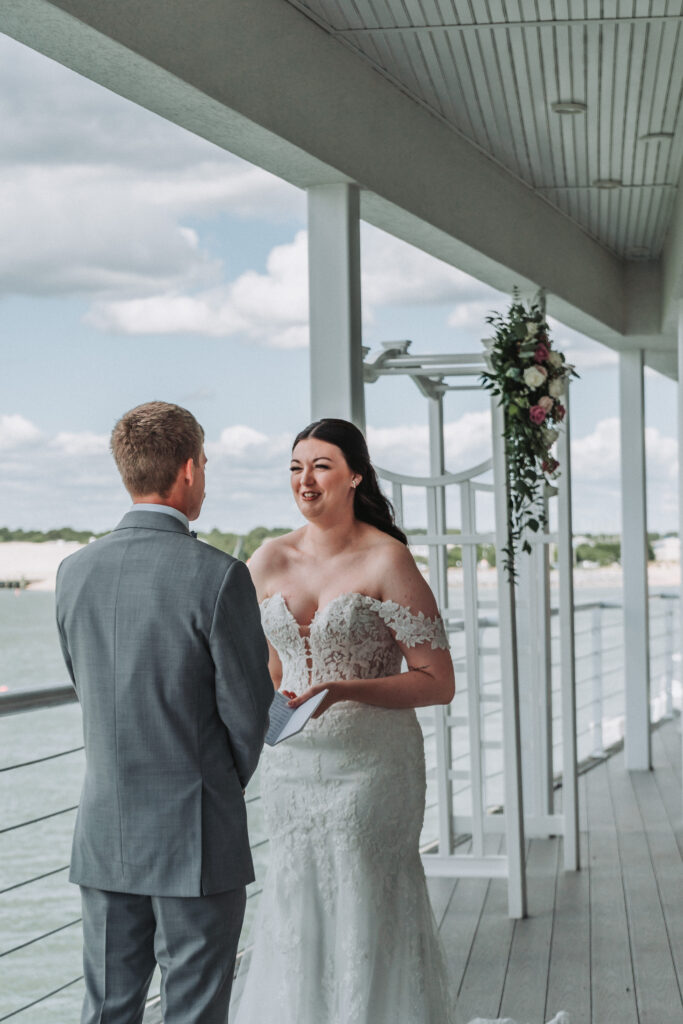 Bride reading her vows to the groom on the deck of the Lesner Inn with a floral arch behind them.