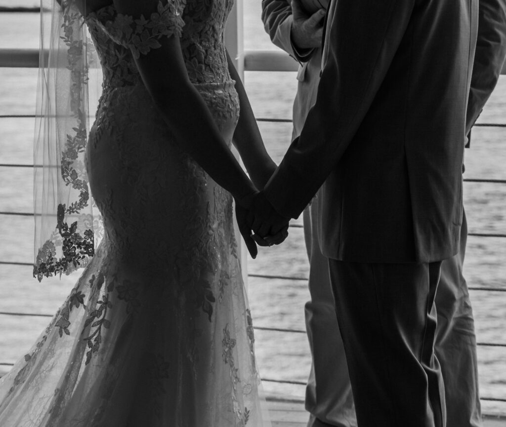 Bride and groom holding hands during their waterfront ceremony at the Lesner Inn, with a focus on their joined hands.