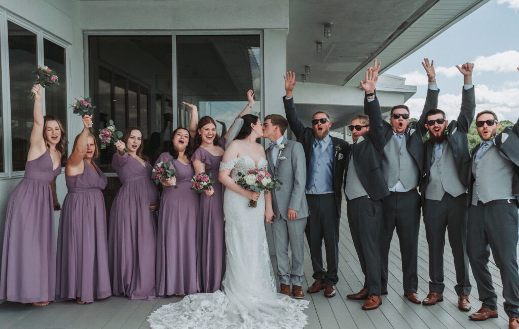 Bride and groom kiss while wedding party in lavender and gray cheer around them at the Lesner Inn.