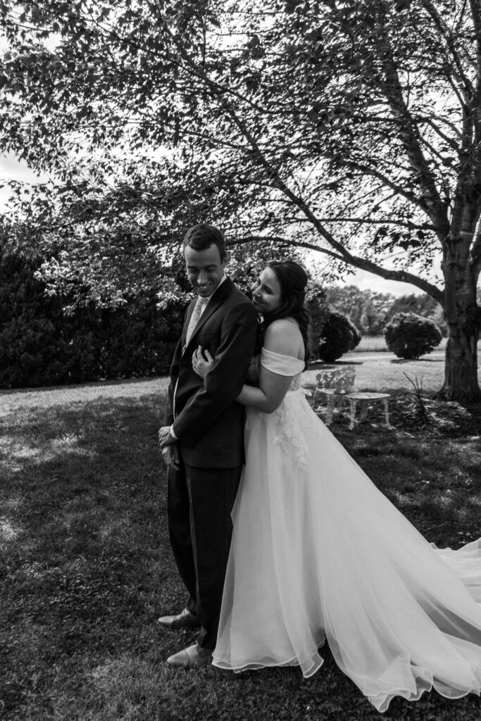Bride hugs groom from behind while laughing in black and white photo at Due Sorelle