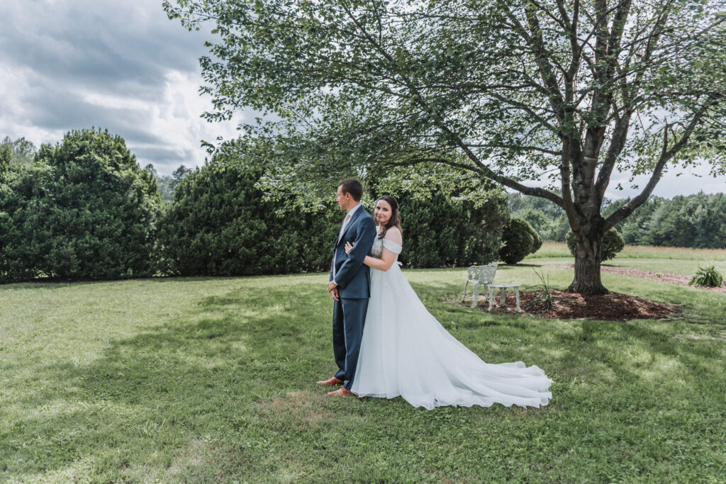 Bride and groom standing in a green garden with large tree and iron bench in background
