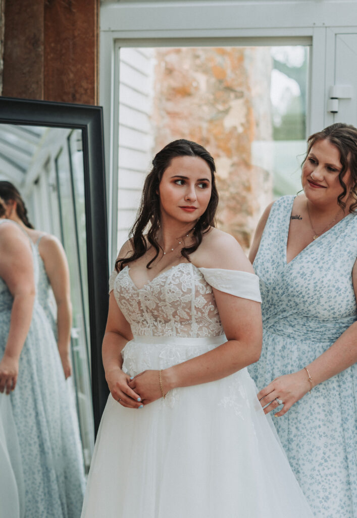 Bride smiling in front of mirror as bridesmaid helps adjust gown at Due Sorelle