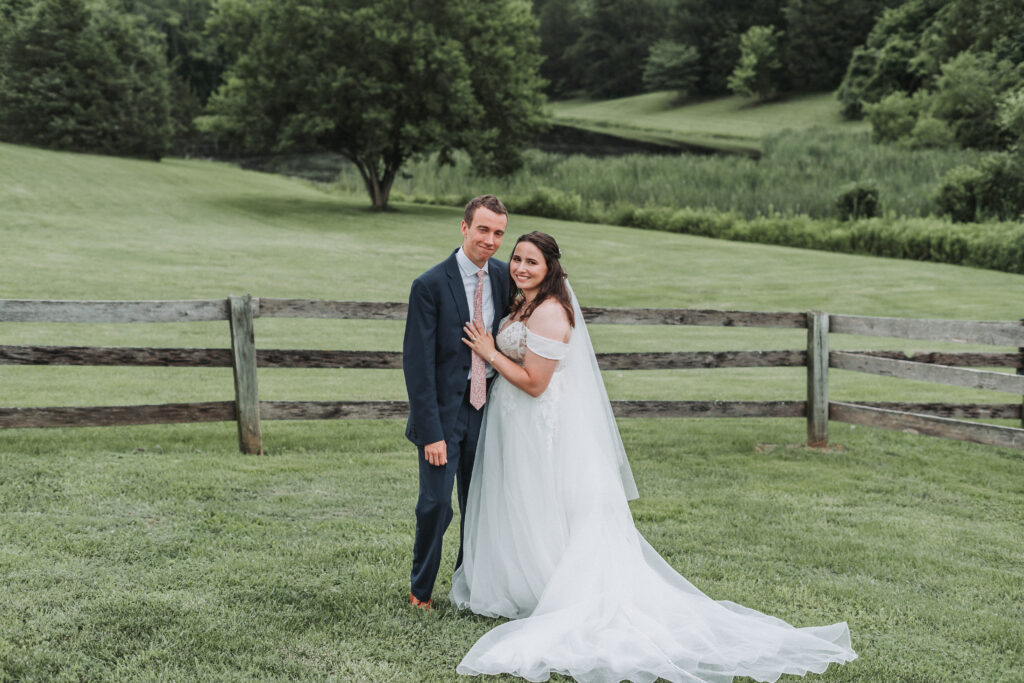 Bride and groom pose with rolling hills behind them and a rustic wooden fence at Due Sorelle in Ruckersville, VA.