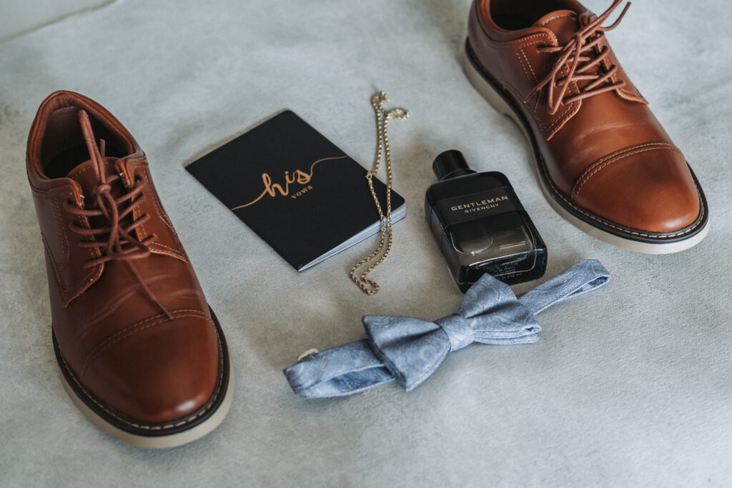 Groom’s flat lay with brown leather shoes, blue bow tie, Gentleman cologne, chain, and vow book.