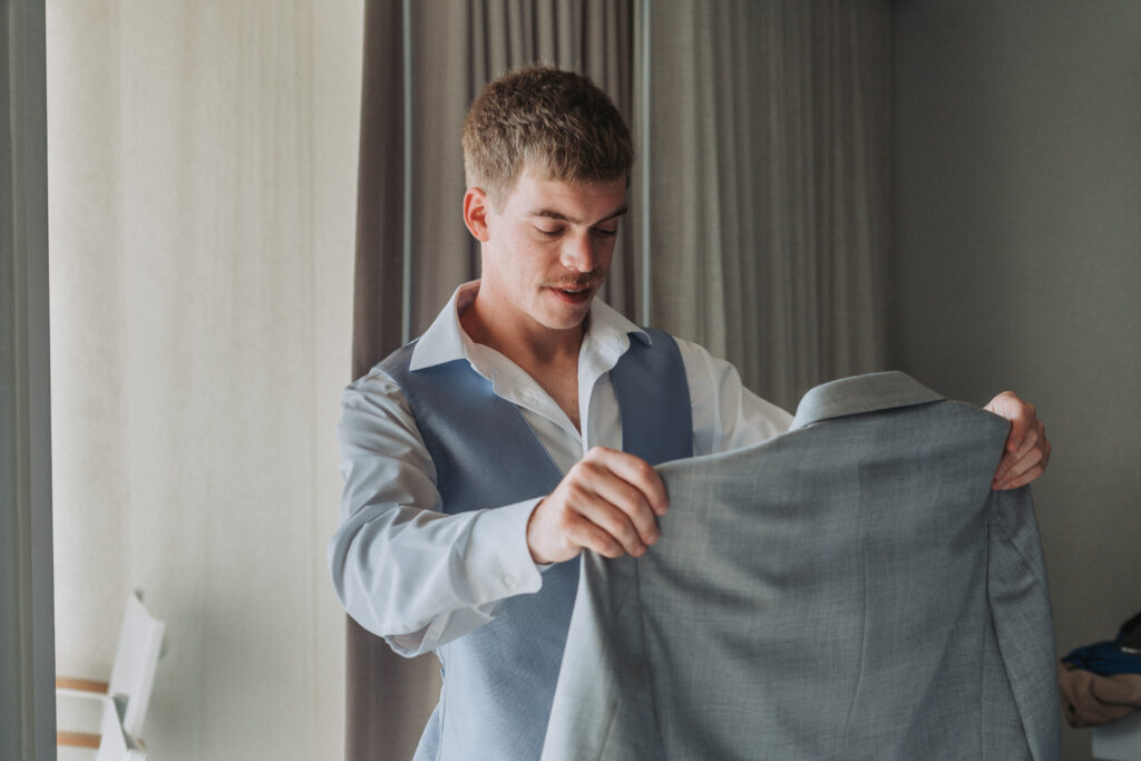 Groom looking down while holding his light gray suit jacket, preparing to get dressed.